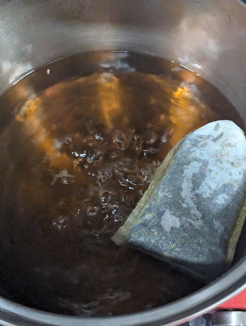 May include: A stainless steel pot filled with boiling, dark brown liquid. A tea bag is partially submerged in the water. Bubbles rise to the surface, indicating the water is hot. The pot sits on a red stovetop.