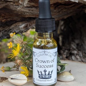 K&ouml;nnte beinhalten: Eine kleine Glasflasche mit schwarzem Tropfer und einem wei&szlig;en Etikett, auf dem "Conjure Oil Crown of Success" mit einer Kronengrafik und "Conjured Moon" unten steht. Die Flasche steht auf einer Holzoberfl&auml;che mit einer Muschel auf jeder Seite.