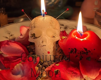 Könnte beinhalten: Zwei Kerzen, eine weiße schädelförmige und eine rote, brennen auf einem Teller mit Rosenblättern und getrockneten Kräutern. Der Text "Conjured Moon" ist auf dem Teller geschrieben.