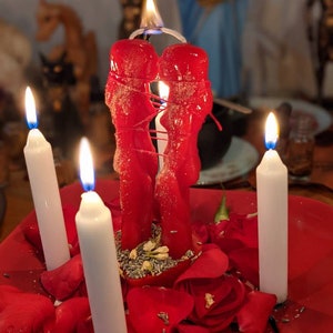 Op de afbeelding: Een rode kaars in de vorm van twee figuren die elkaar omhelzen, omringd door witte kaarsen en rode rozenblaadjes op een rood bord.