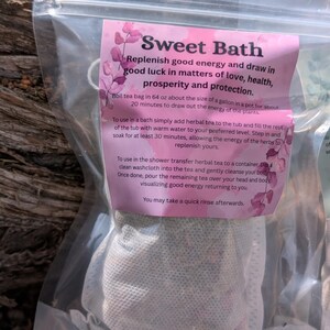 May include: A clear plastic bag containing a tea bag labeled "Sweet Bath." The pink label includes instructions for use and the phrase "Replenish good energy and draw in good luck." The tea bag is white and appears to be filled with herbs.