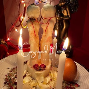 May include: Two white candles shaped like figures of a couple, standing on a white base with rose petals and herbs. The candles are lit and there are three white candles surrounding them. The base has the text "Conjured Moon Botanica" on it. There is a brown egg in the background.
