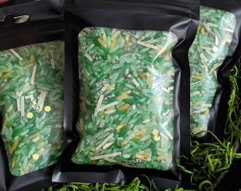 May include: Three black zippered pouches filled with green and yellow rice, resembling money. The pouches are sitting on a bed of green shredded paper.