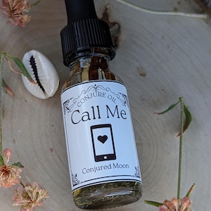 May include: A small glass bottle with a black dropper top filled with a clear liquid and herbs. The label reads "Conjure Oil Call Me" with a phone icon and a heart. The label also says "Conjured Moon".