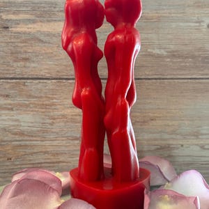 May include: A red candle shaped like two figures kissing. The candle is on a heart-shaped base and is surrounded by pink rose petals.