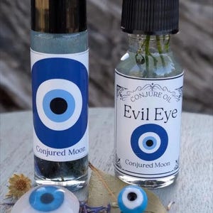 May include: Two glass bottles with black caps and labels featuring a blue and white evil eye design. One bottle is a rollerball, the other has a dropper. The text "Conjured Moon" and "Evil Eye" are visible. A small evil eye bead and a white evil eye charm are also present.