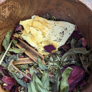 May include: A wooden bowl filled with a variety of dried herbs and botanicals. The mix includes green leaves, dried petals in shades of red and purple, and a large piece of dried fruit. A cinnamon stick is also visible.