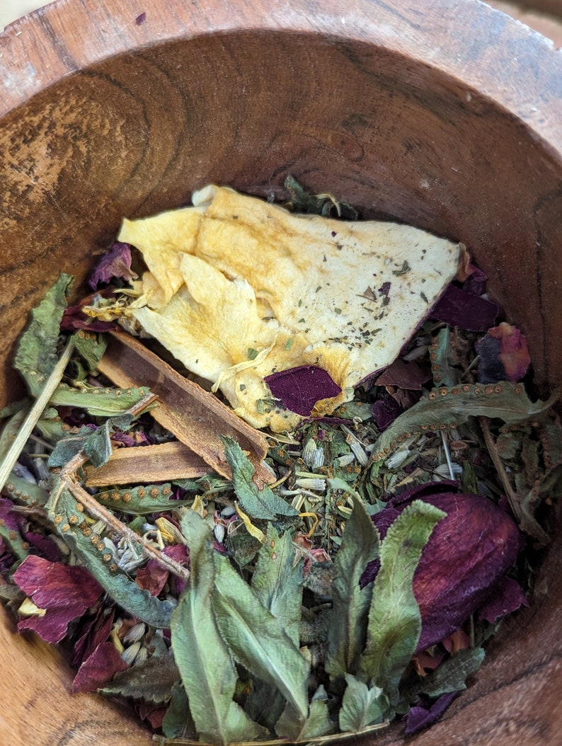 May include: A wooden bowl filled with a variety of dried herbs and botanicals. The mix includes green leaves, dried petals in shades of red and purple, and a large piece of dried fruit. A cinnamon stick is also visible.