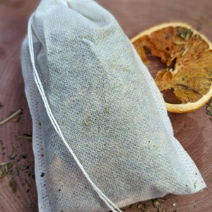 May include: A white tea bag filled with dried herbs and spices, tied with a white string. A dried orange slice sits to the right of the tea bag on a wooden surface. Scattered herbs are visible around the tea bag and orange slice.