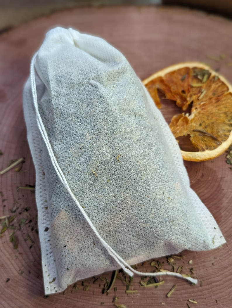 May include: A white tea bag filled with dried herbs and spices, tied with a white string. A dried orange slice sits to the right of the tea bag on a wooden surface. Scattered herbs are visible around the tea bag and orange slice.