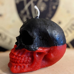 May include: A black and red skull-shaped candle with a white wick. The skull is painted black on the top half and red on the bottom half.