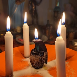 May include: Five white candles are lit and arranged in a circle around a black grenade-shaped candle holder on an orange plate. The candles are on a white surface with a white powder sprinkled around them.