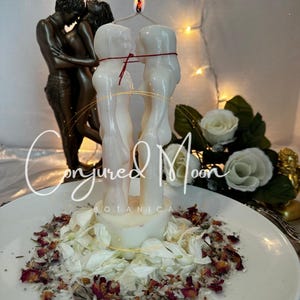 May include: White candle with two figures embracing, tied with red string, on a white plate with white flower petals and red rose petals. The candle is lit and the text "Conjured Moon Botanica" is visible below the candle.