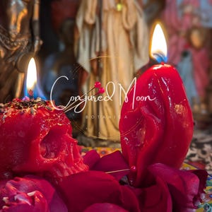 Puede incluir: Dos velas rojas, una con forma de calavera y la otra una vela tradicional, están ardiendo en un plato con pétalos de rosa. Las velas están rodeadas de hierbas secas y un pequeño alfiler. El texto "Conjured Moon Botanica" es visible en el fondo.