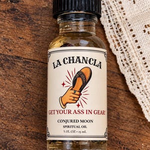 May include: A clear glass bottle of spiritual oil with a black cap and a cream-colored label. The label features the text LA CHANCLA, GET YOUR ASS IN GEAR!, CONJURED MOON, SPIRITUAL OIL, and 1/2 FL. OZ. 15 mL, with an illustration of a hand holding a sandal.