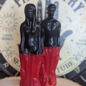May include: Two black and red candles shaped like human figures. The candles are standing upright on a wooden surface.