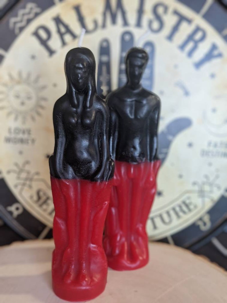 Op de afbeelding: Twee zwarte en rode kaarsen in de vorm van menselijke figuren. De kaarsen staan rechtop op een houten oppervlak.