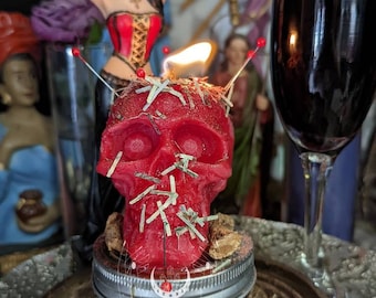 Könnte beinhalten: Eine rote Schädel-Kerze mit einer Flamme, die oben brennt. Die Kerze ist mit roten Stecknadeln und kleinen Papierstücken bedeckt. Die Kerze steht in einem Glasgefäß mit einem silbernen Deckel. Das Gefäß trägt die Aufschrift "CONJURED MOON" auf der Vorderseite.
