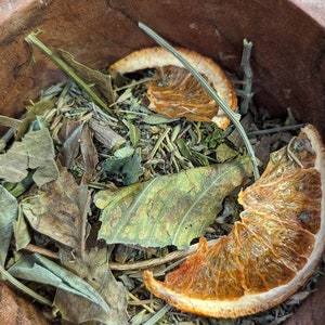 May include: A wooden bowl filled with dried herbs and orange slices. The herbs are a mix of green and brown leaves and stems. The orange slices are a vibrant orange color and add a pop of color to the arrangement. The bowl is made of a dark wood.