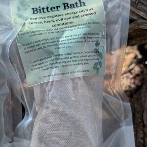 May include: A clear plastic bag containing a tea bag labeled "Bitter Bath." The label, with a green and white design, details instructions for removing negative energy. The tea bag is filled with dried herbs.