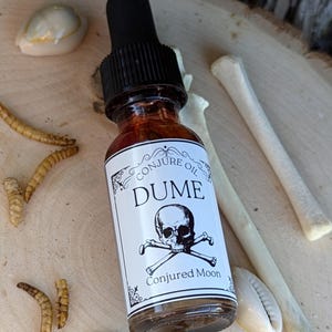 May include: A small glass bottle with a black dropper top and a white label that reads "Conjure Oil DUME" with a skull and crossbones graphic and the text "Conjured Moon".