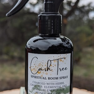 May include: A black glass bottle with a spray nozzle, labeled "Cash Tree Spiritual Room Spray". The bottle is sitting on a pile of US dollar bills and green stones. The label says "Charged with herbs, crystals elemental and spiritual energy for money, success, abundance and luck. Conjurer Moon 6 oz/177mL."