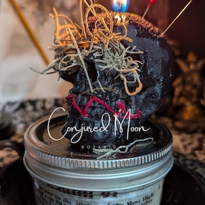 Pode incluir: Uma vela preta em forma de caveira com alfinetes vermelhos e musgo, sentada em um frasco de vidro com tampa prateada. O frasco tem um rótulo que diz "Conjured Moon Botanics" e uma passagem de texto sobre a "sombra do Todo-Poderoso".