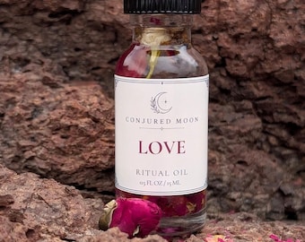 May include: A clear glass bottle with a black cap containing ritual oil and dried rose petals. The white label features the text CONJURED MOON, LOVE, RITUAL OIL, and 0.5 FL OZ/15 ML. The bottle is filled with liquid and botanical elements.
