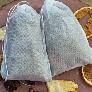 May include: Two white fabric tea bags filled with dried herbs and flowers, tied with a white string. The bags are on a wooden surface with dried orange slices and other botanicals.