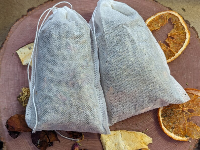 May include: Two white fabric tea bags filled with dried herbs and flowers, tied with a white string. The bags are on a wooden surface with dried orange slices and other botanicals.