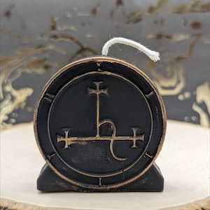 May include: A black candle with a bronze-colored emblem featuring a cross and other symbols. The candle has a white wick and sits on a wooden surface. The candle is round and has a distressed finish.