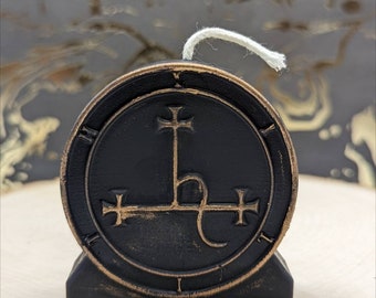 May include: A black candle with a bronze-colored emblem featuring a cross and other symbols. The candle has a white wick and sits on a wooden surface. The candle is round and has a distressed finish.