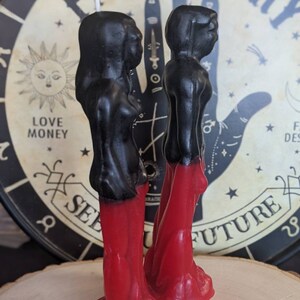 Op de afbeelding: Twee zwarte en rode kaarsfiguren die naast elkaar staan. De figuren zijn gekleed in lange rode gewaden en hebben zwarte hoofden en schouders. De kaarsen staan op een houten oppervlak voor een zwart-witte achtergrond met de woorden "Palmistry See Your Future".