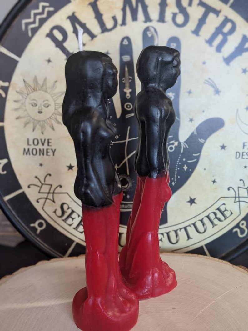 Op de afbeelding: Twee zwarte en rode kaarsfiguren die naast elkaar staan. De figuren zijn gekleed in lange rode gewaden en hebben zwarte hoofden en schouders. De kaarsen staan op een houten oppervlak voor een zwart-witte achtergrond met de woorden "Palmistry See Your Future".