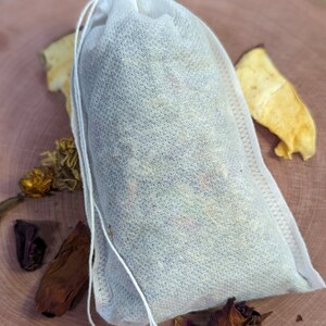 May include: A white tea bag filled with dried herbs and flowers, tied with a white string. The tea bag is on a wooden surface, with dried apple slices and other botanicals scattered around it. The tea bag is filled with a variety of colorful dried herbs.