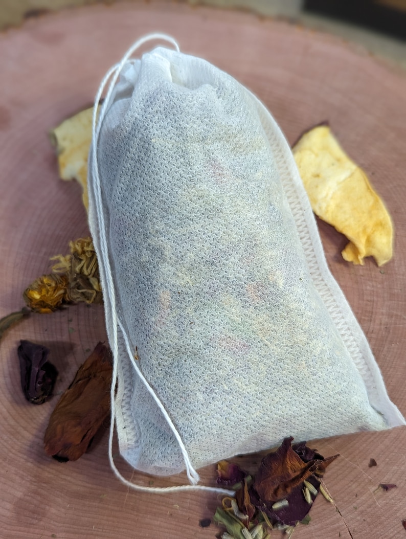 May include: A white tea bag filled with dried herbs and flowers, tied with a white string. The tea bag is on a wooden surface, with dried apple slices and other botanicals scattered around it. The tea bag is filled with a variety of colorful dried herbs.