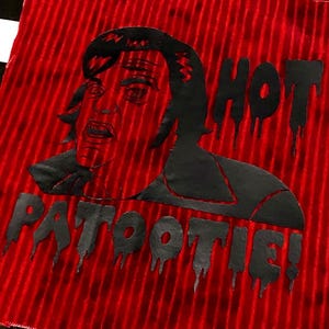 Puede incluir: Un parche de tela roja texturizada con un gráfico negro de la cara de una persona y las palabras "HOT PATOOTIE!" en una fuente goteante. El parche es cuadrado y tiene un borde sin rematar.