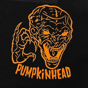 May include: Black square patch featuring an orange illustration of a monster with sharp claws and teeth. The text "PUMPKINHEAD" is printed in orange below the monster. The patch is on a black and white checkered surface.