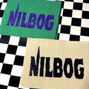 May include: Two rectangular patches with the word "NILBOG" printed on them. One patch is green with purple text, and the other is tan with black text. The patches are on a black and white checkered surface.