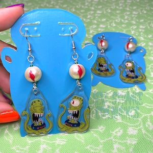 Könnte beinhalten: Ohrringe mit Alien-Motiv zum Aufhängen. Jeder Ohrring zeigt einen Cartoon-Alien in einer blauen Weltraumkapsel, mit einer cremefarbenen Perle mit rotem Streifen und einer grünen Perle. Die Ohrringe werden auf einer blauen Karte präsentiert.