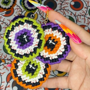 May include: A pair of handmade Halloween-themed earrings featuring a colorful eye design with black, white, orange, green, and purple yarn. The earrings are crocheted and have a dangling design.