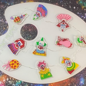 May include: A string of fairy lights with clown faces on each bulb. The lights are strung on a white background that resembles a painter's palette. The clowns are all different colours and have different expressions.