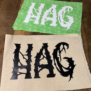 May include: Two fabric patches with the word "HAG" in a gothic font. One patch is light green with a floral pattern and white lettering. The other is beige with black lettering. The patches are likely for sewing or crafting.