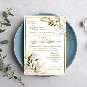 May include: A wedding invitation with gold accents, featuring the names Lorena & Mauricio. The invitation includes floral designs and text in Spanish, with the date, time, and location details. The invitation is on a blue plate.