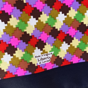 May include: A fabric textile featuring a vibrant, multi-colored houndstooth pattern in shades of red, purple, green, brown, and cream. A white triangular label is printed with the text Made in England Liberty of London. A solid navy blue border.