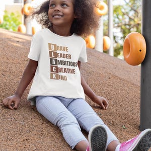 May include: A young person wearing a white t-shirt with the words "BLACK" spelled vertically with each letter representing a positive attribute: "Brave", "Leader", "Ambitious", "Creative", and "Kind".