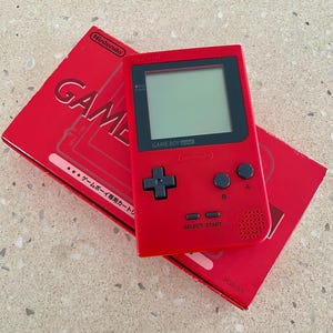 K&ouml;nnte beinhalten: Ein roter Game Boy Pocket Handheld-Videospielkonsole mit seiner roten Box. Die Konsole hat einen schwarzen Bildschirm und schwarze Kn&ouml;pfe. Auf der Box steht das Wort "GAME".