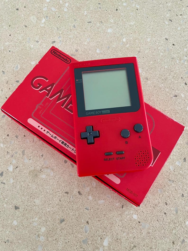 K&ouml;nnte beinhalten: Ein roter Game Boy Pocket Handheld-Videospielkonsole mit seiner roten Box. Die Konsole hat einen schwarzen Bildschirm und schwarze Kn&ouml;pfe. Auf der Box steht das Wort "GAME".