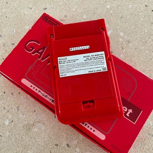 K&ouml;nnte beinhalten: Eine rote Nintendo Game Boy Konsole und ihre Box werden gezeigt. Auf der R&uuml;ckseite der Konsole ist die Modellnummer MGB-001 und weitere Details zu sehen. Auf der Box ist das Wort "Game" sichtbar. Konsole und Box liegen auf einer hellen Oberfl&auml;che.