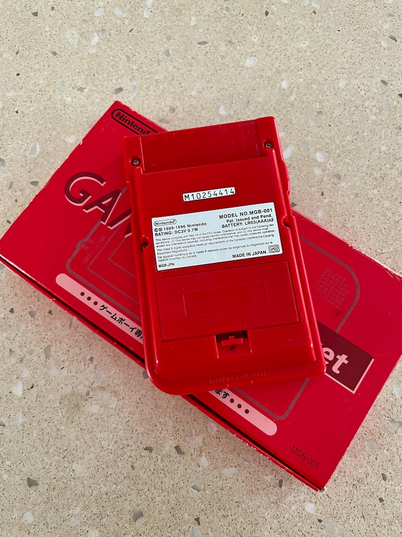 K&ouml;nnte beinhalten: Eine rote Nintendo Game Boy Konsole und ihre Box werden gezeigt. Auf der R&uuml;ckseite der Konsole ist die Modellnummer MGB-001 und weitere Details zu sehen. Auf der Box ist das Wort "Game" sichtbar. Konsole und Box liegen auf einer hellen Oberfl&auml;che.